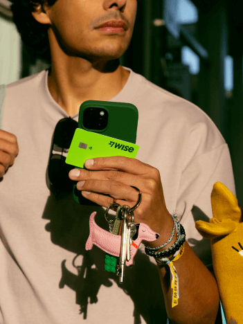 Un homme marche dans une rue commerçante. Il tient son téléphone, ses clés, ses lunettes de soleil et une carte Wise dans sa main.